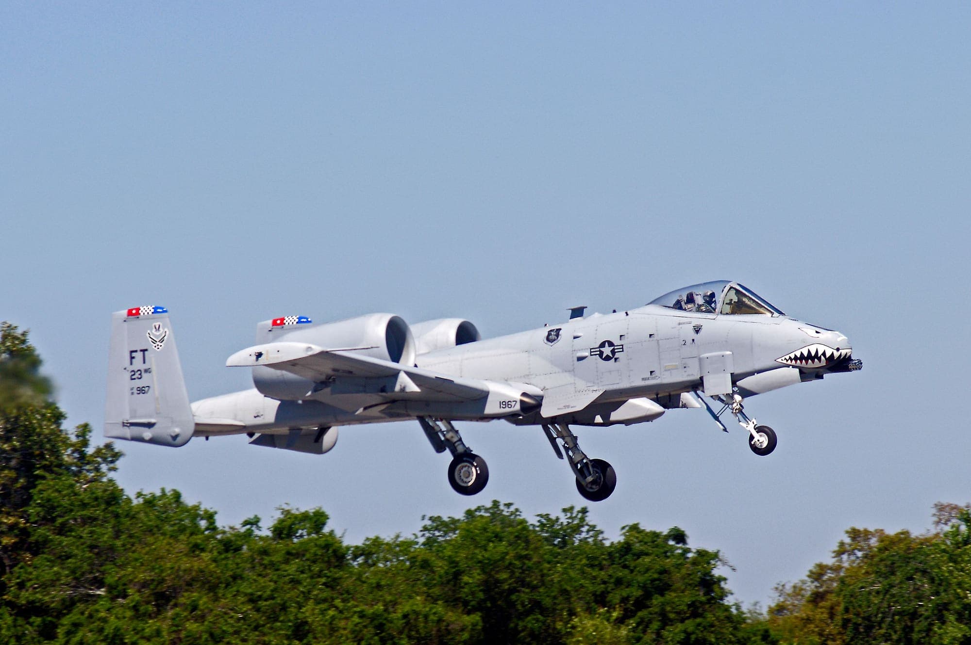 A gray A-10 Thunderbolt II attack jet flying low over a line of green treetops