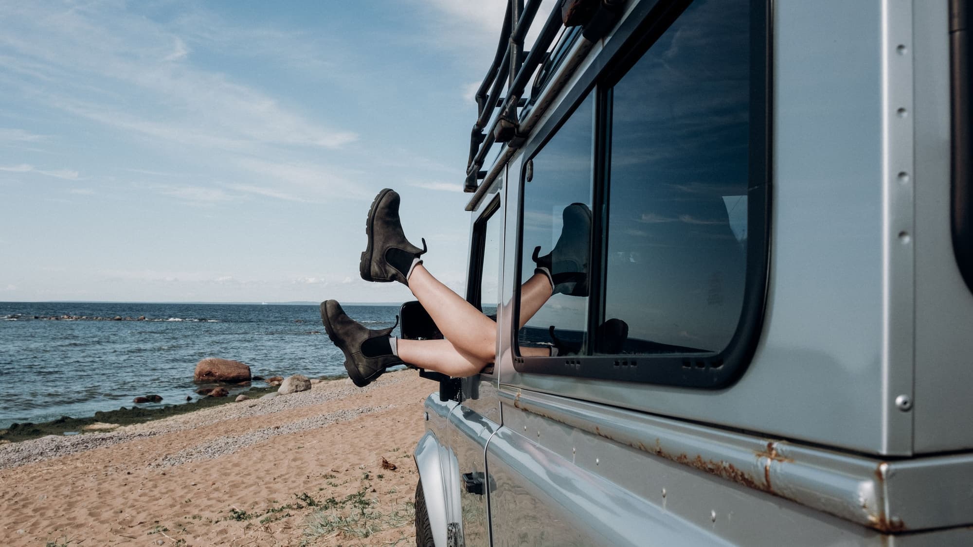 A person's booted feet sticking out of a car window, with a sandy beach and calm water behind