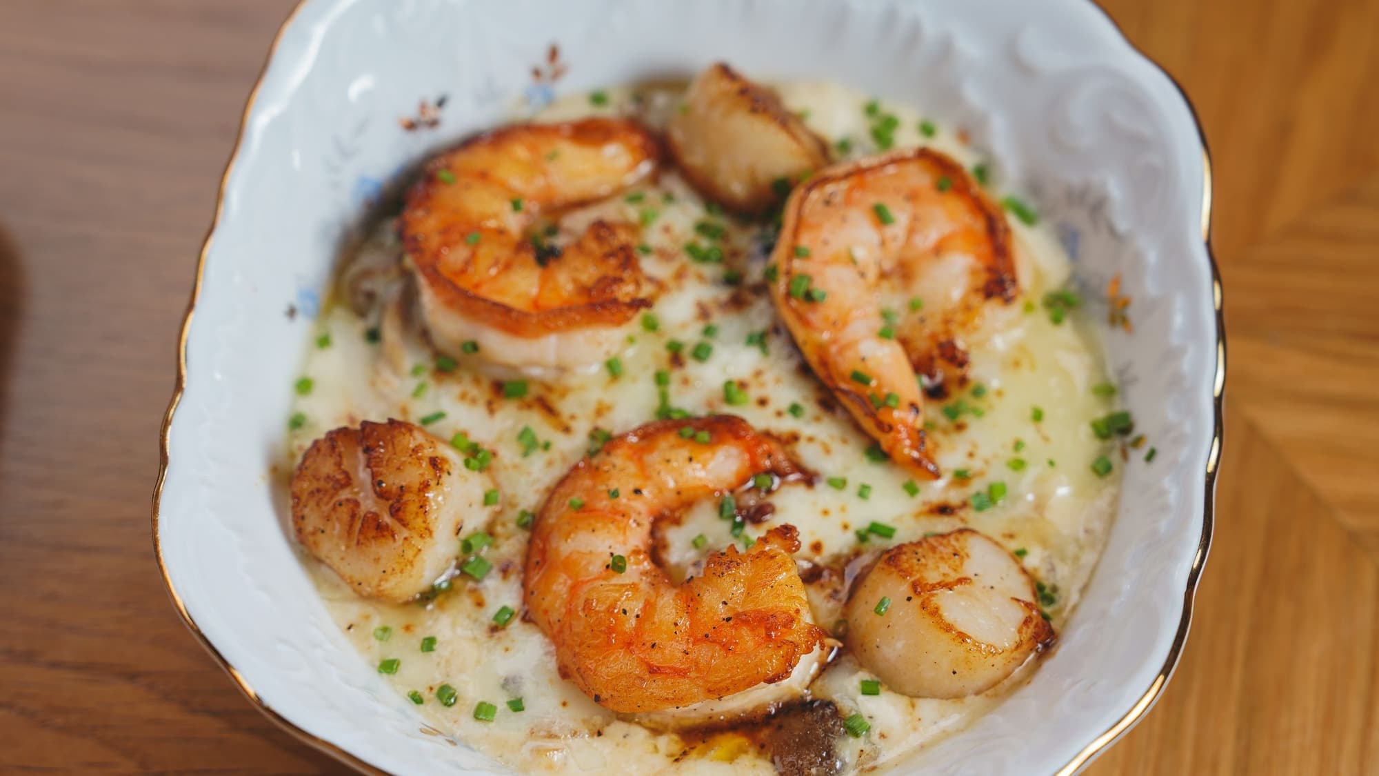 Seared shrimp and scallops over creamy grits in a white porcelain dish, sprinkled with chives
