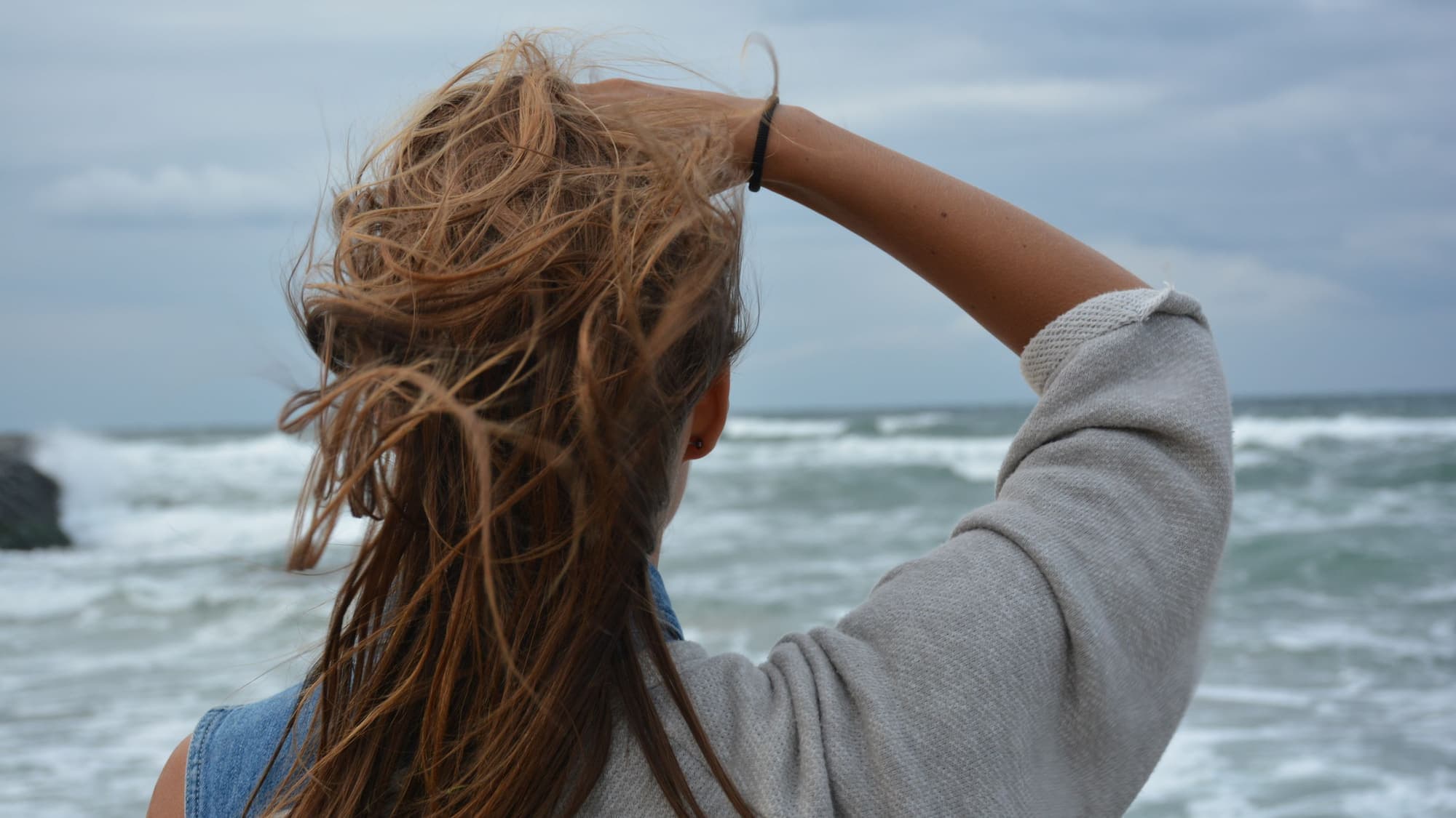 A woman seen from behind at the beach, holding her windblown, wavy hair back from her face