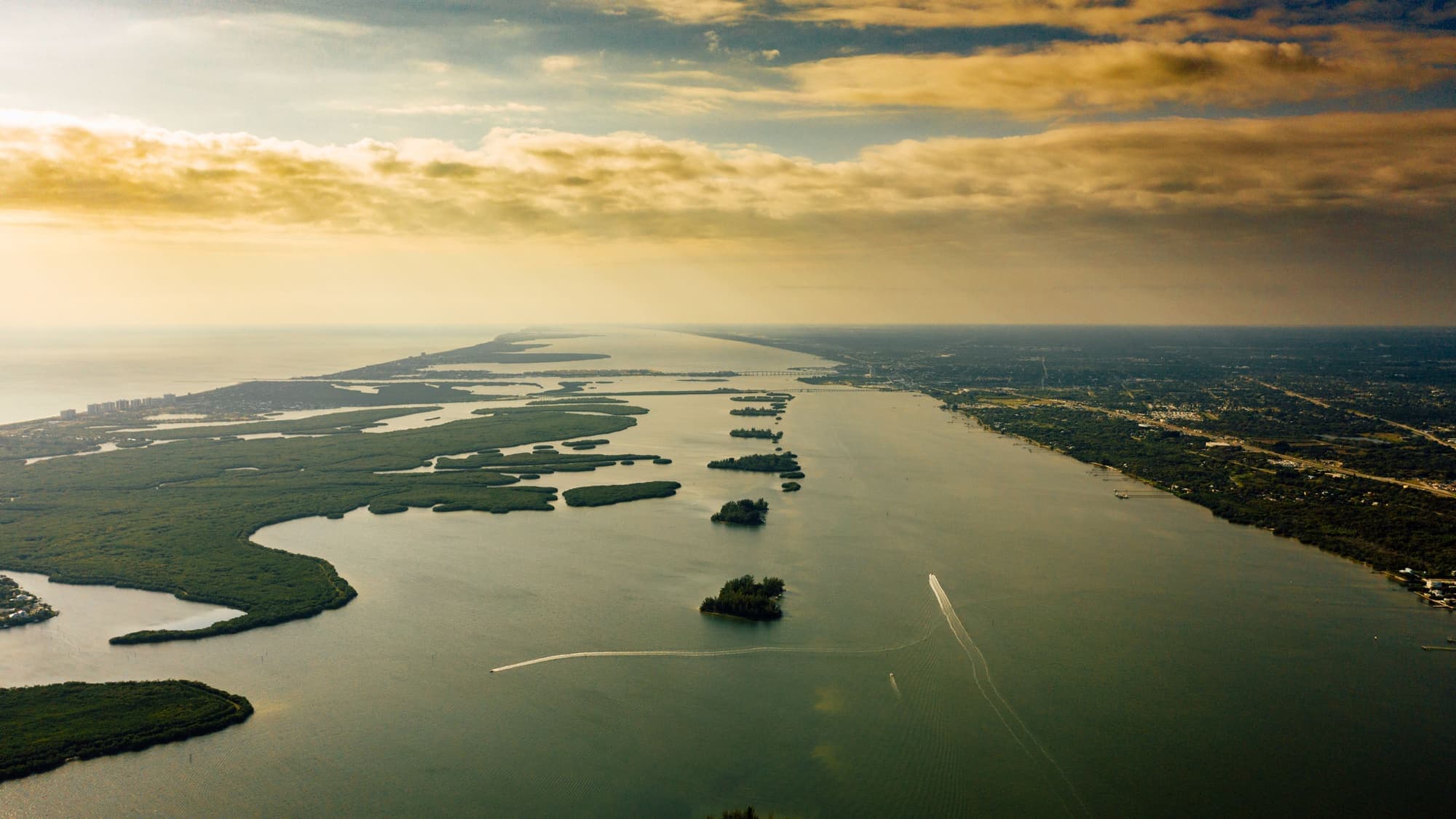 Aerial view of a wide, calm waterway winding between marsh islands under a golden sunset