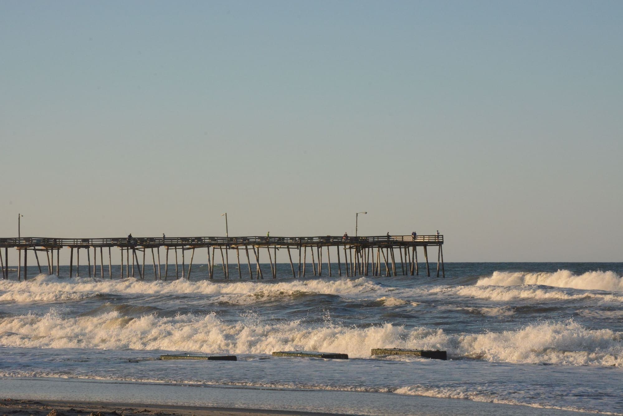 A long wooden fishing pier running out past rolling surf under a soft evening sky