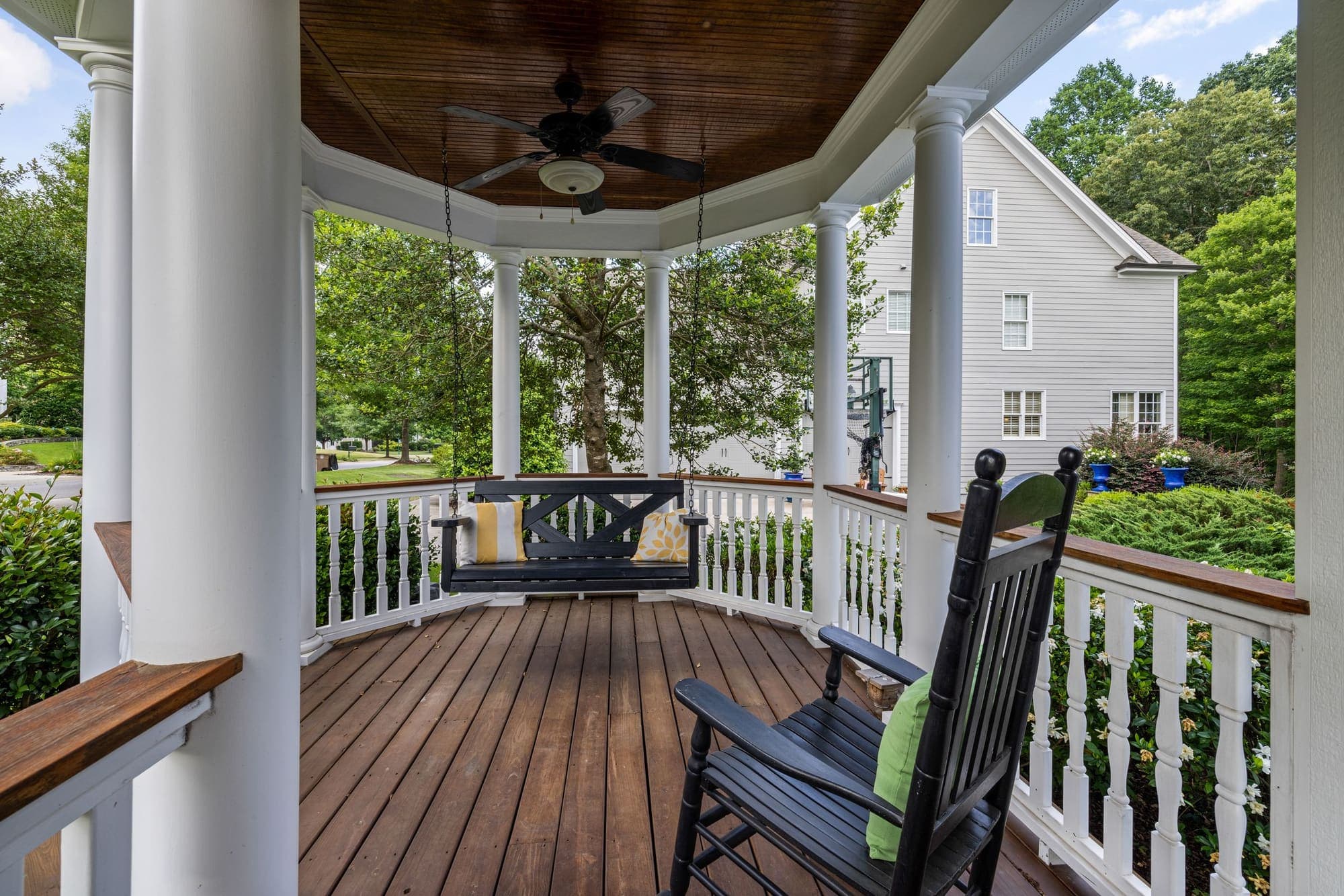 A shaded wraparound porch with white railings, a rocking chair and a porch swing looking out over a green yard
