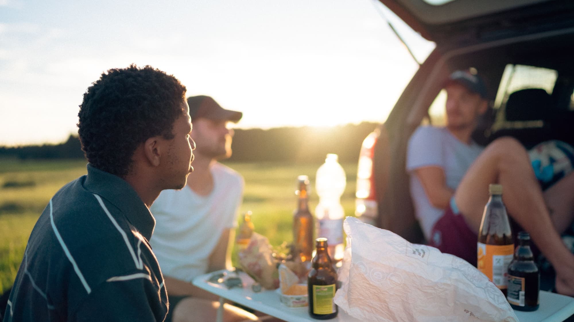Friends relaxing around the open back of a car at sunset with drinks and snacks, tailgate style
