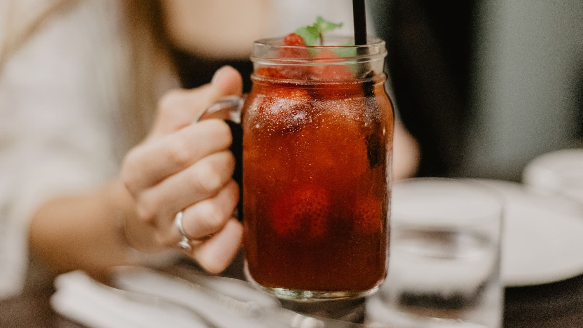 A hand holding a mason jar mug of iced tea with a straw