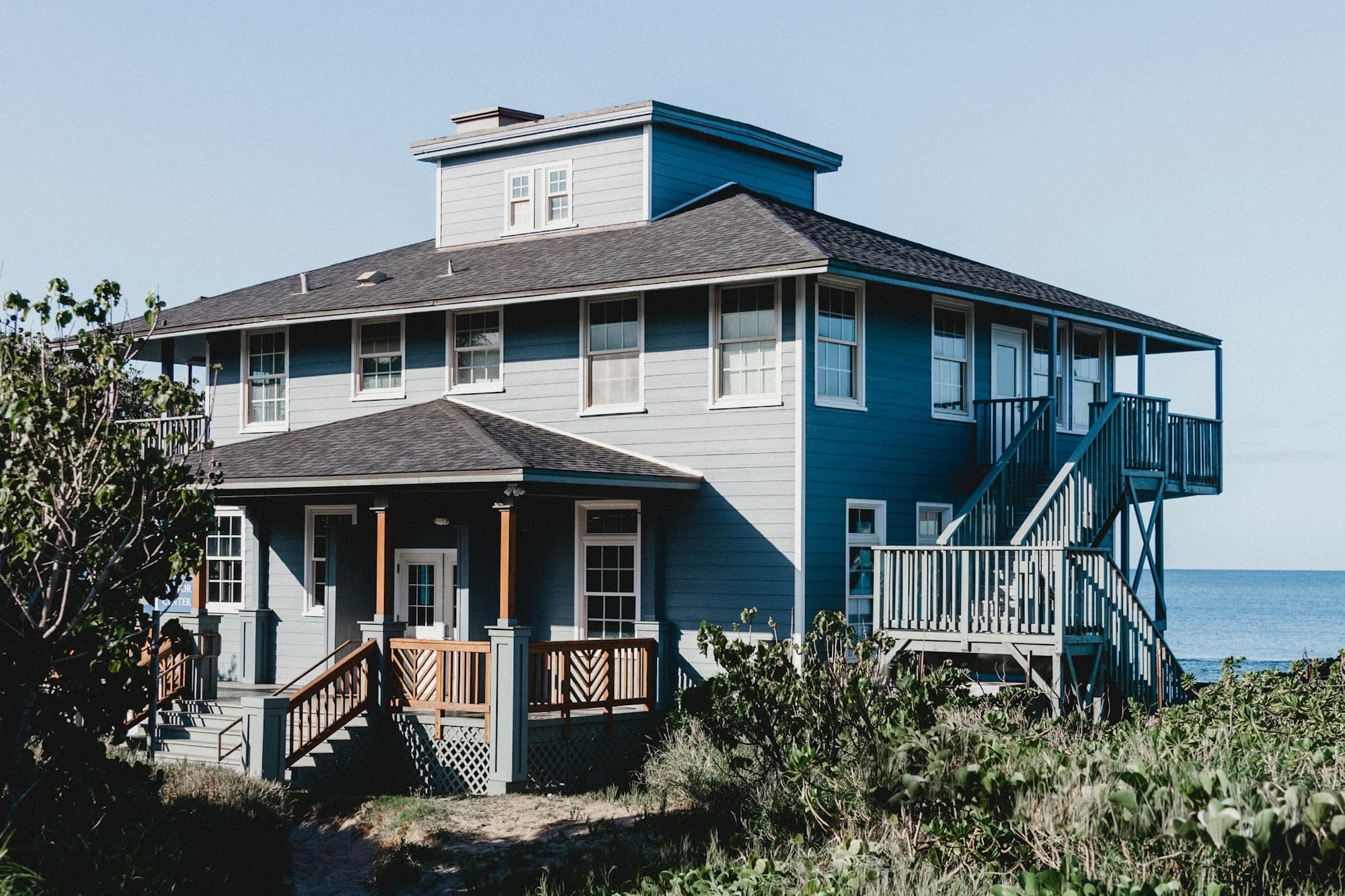 A blue and gray two-story beach house with wraparound porches and exterior stairs, the ocean beyond