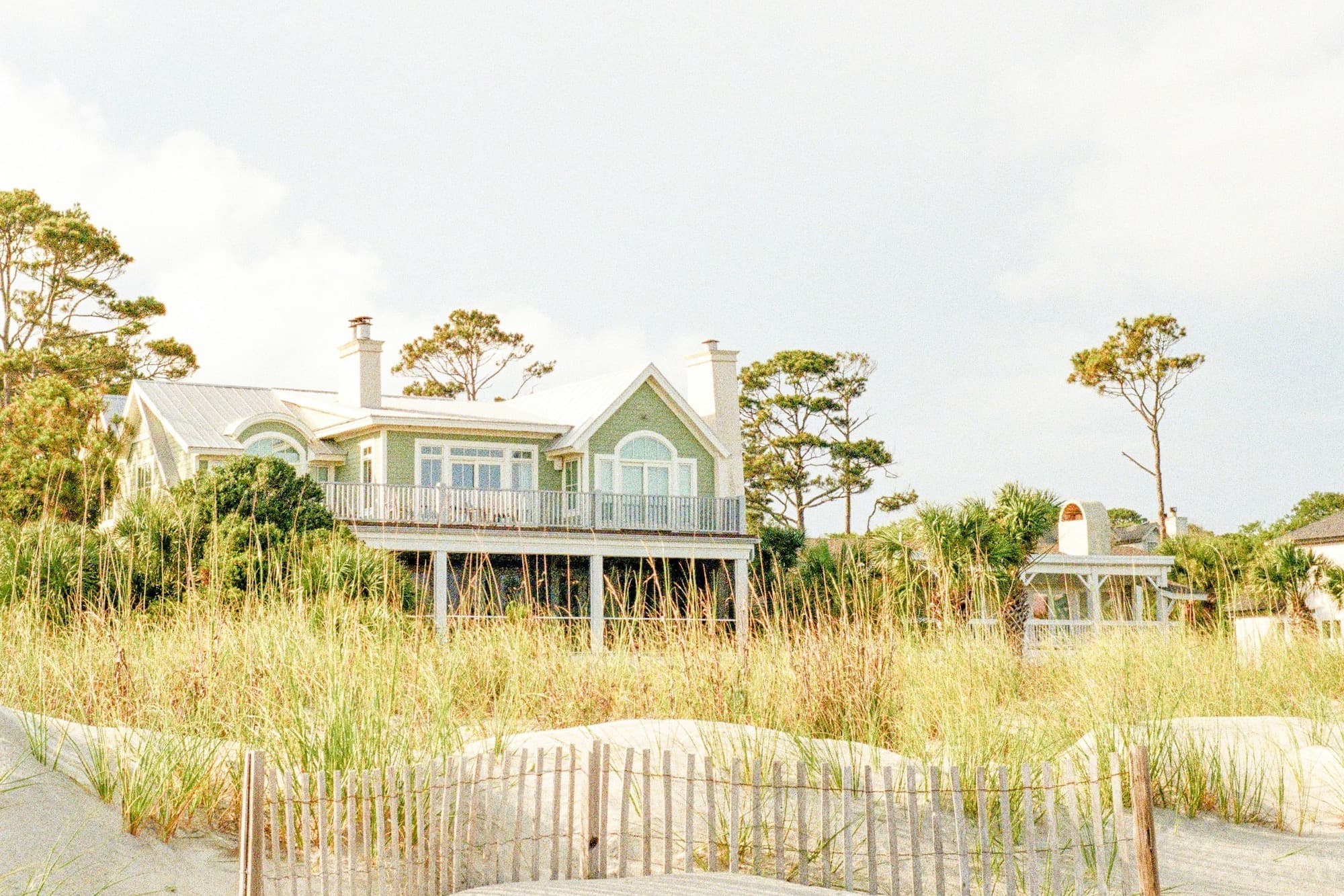 A green two-story beach house raised on pilings behind a dune fence and sea oats