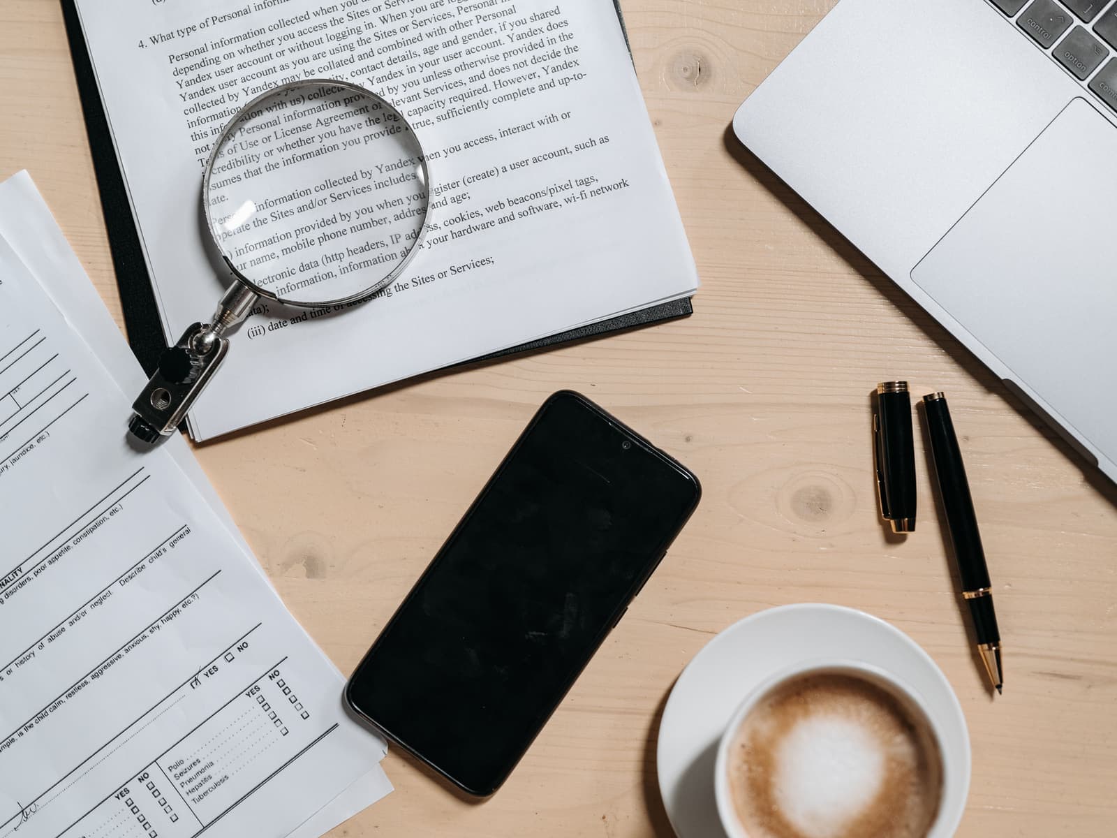 Contract pages, a magnifying glass, pens and a coffee laid out beside a laptop