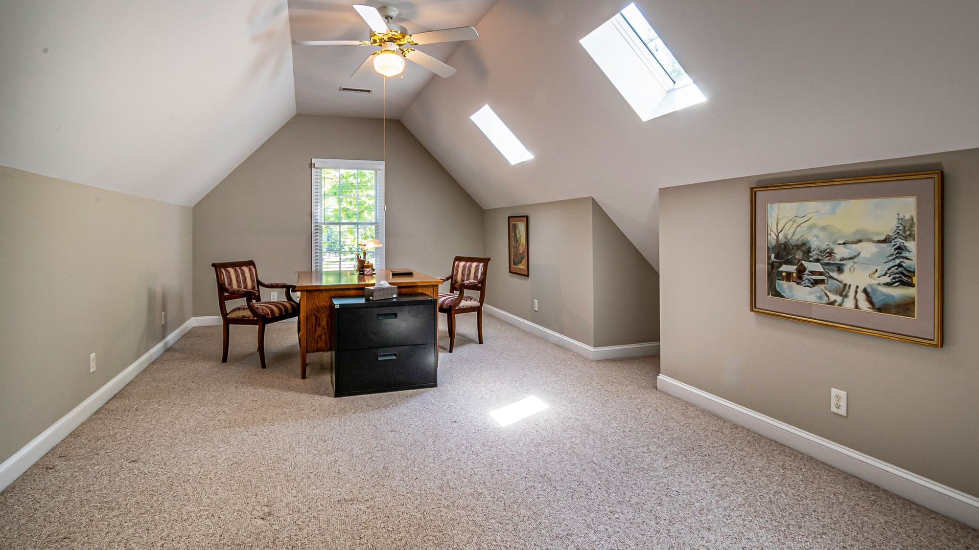 A bright, carpeted bonus room with a sloped ceiling, skylights and a desk by the window
