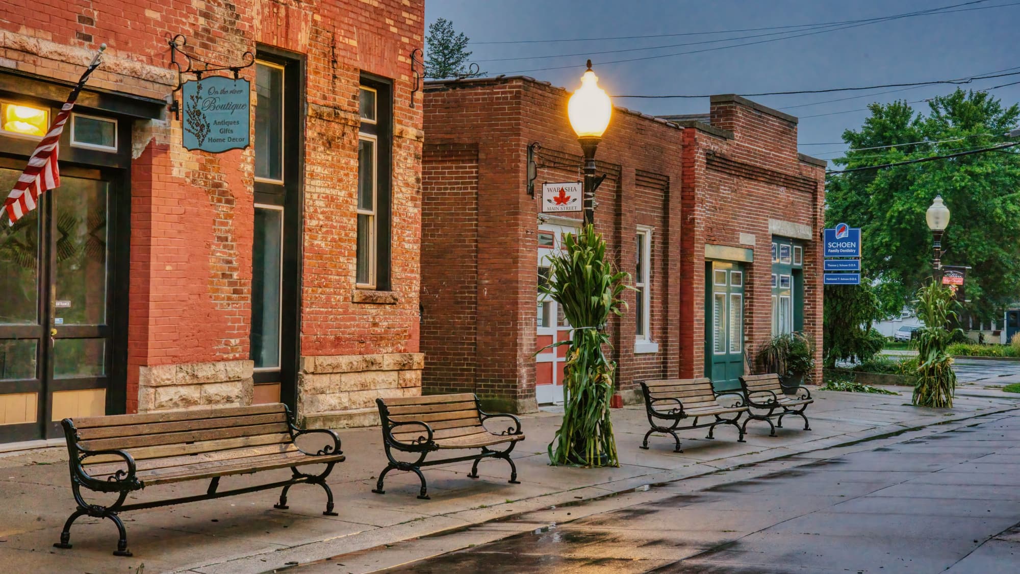 Red brick storefronts and wooden benches along a quiet small-town sidewalk with an old-fashioned street lamp