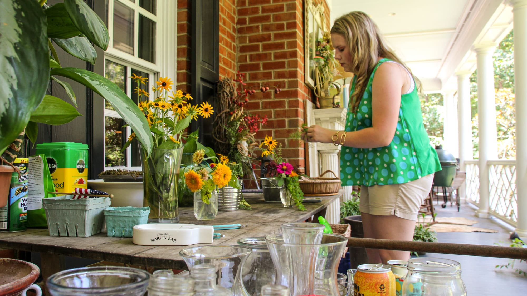 A woman in a green top arranging potted flowers on a covered porch filled with plants