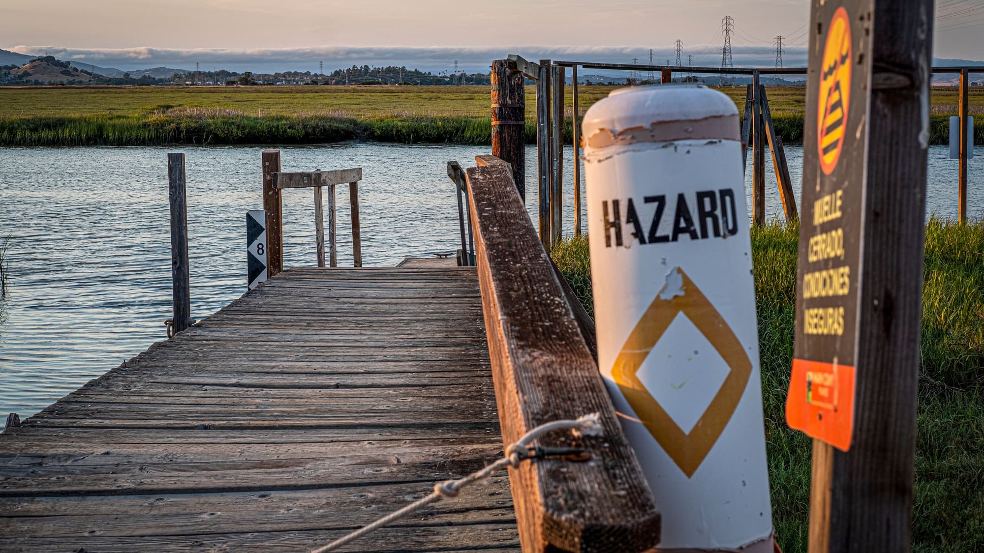 A weathered wooden dock beside a white marker post reading Hazard, with marsh and calm water beyond