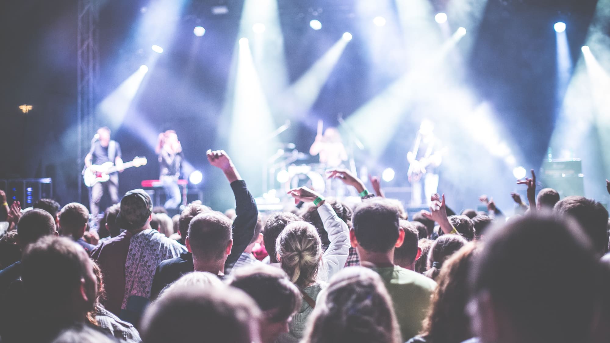 A packed crowd with hands in the air in front of a stage flooded with bright spotlights at night