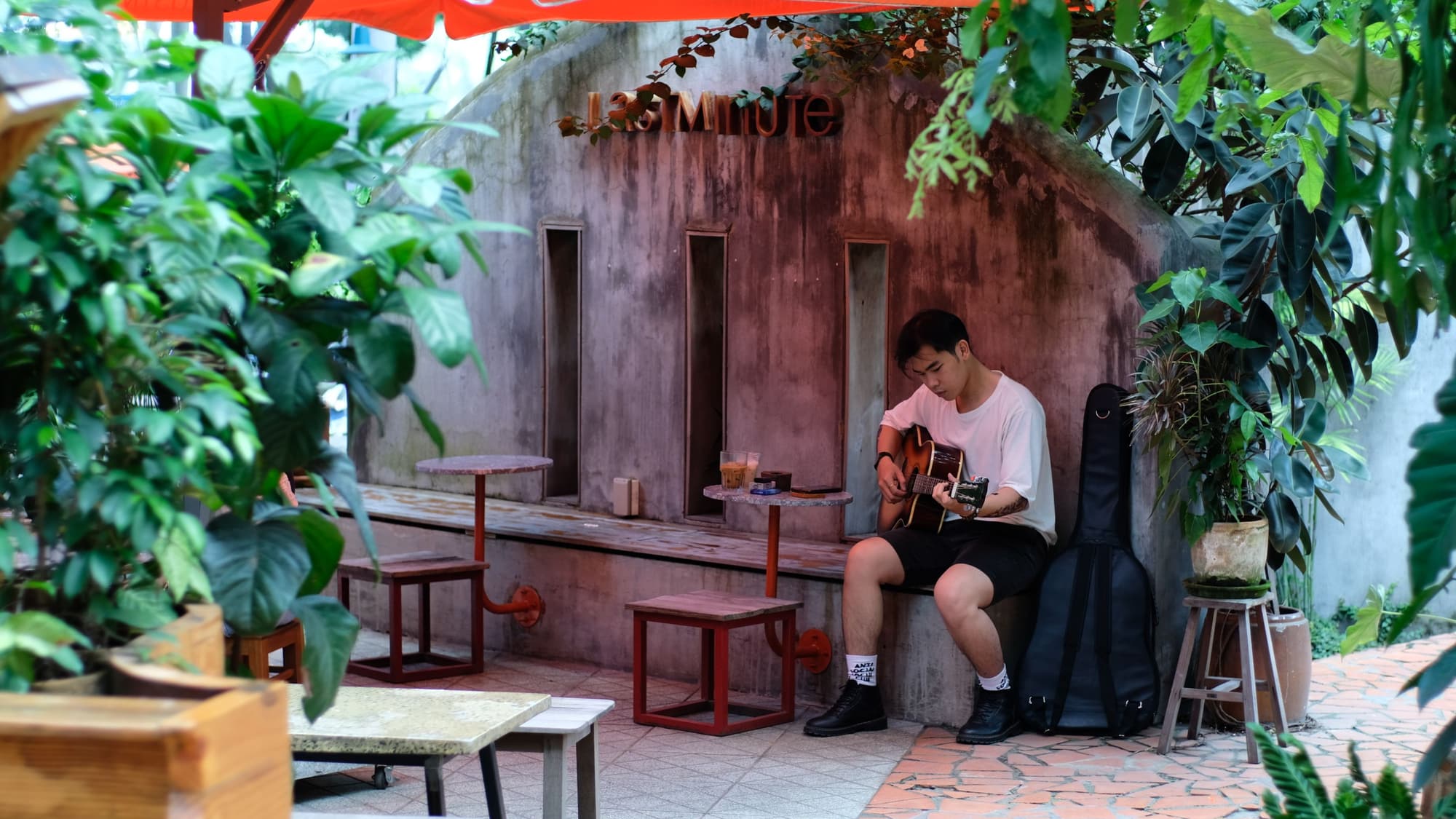 A man sitting on a bench playing an acoustic guitar in a small, leafy courtyard with empty tables around him