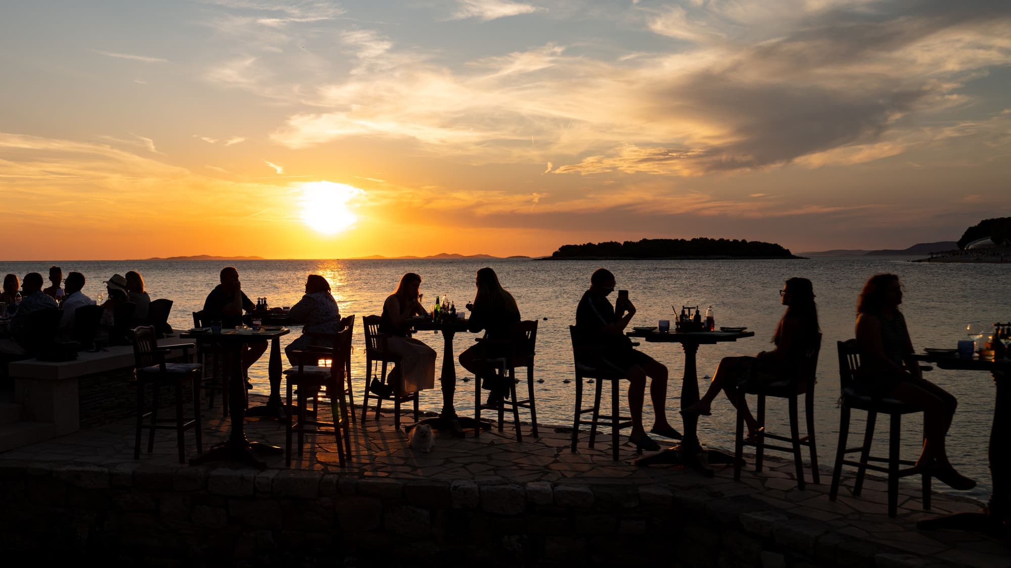 People sitting at high-top tables on a waterfront deck, silhouetted against a sunset over the water