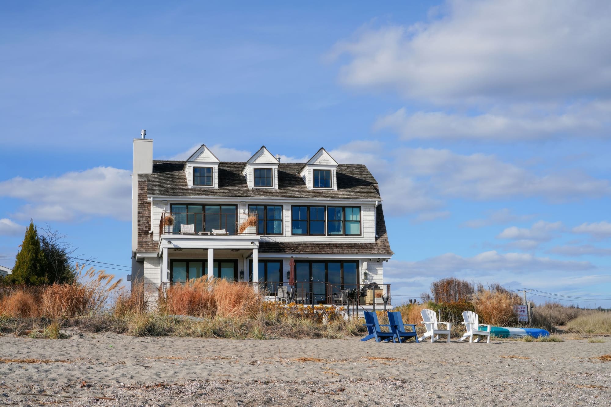 A shingled beach house with dormer windows and patio chairs set out on the sand in front of it