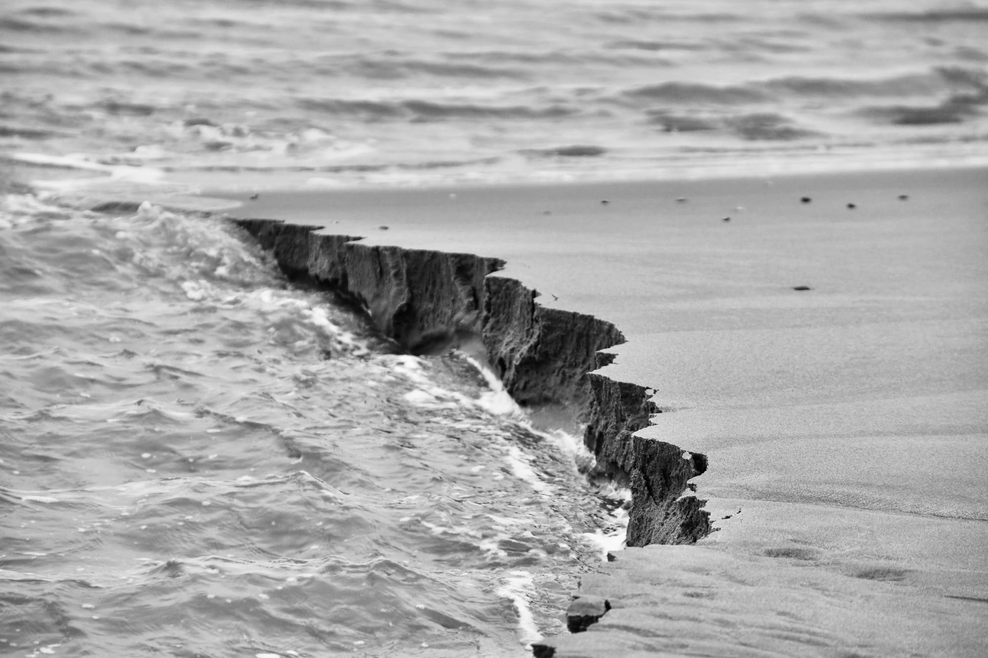 A steep little cliff of sand cut into a beach by the surf, in black and white