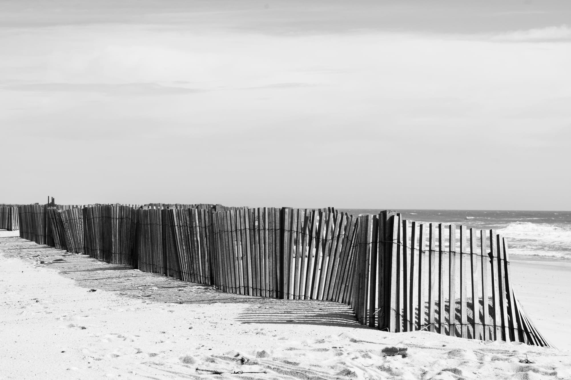 A low sand fence running along a wide, empty beach in black and white