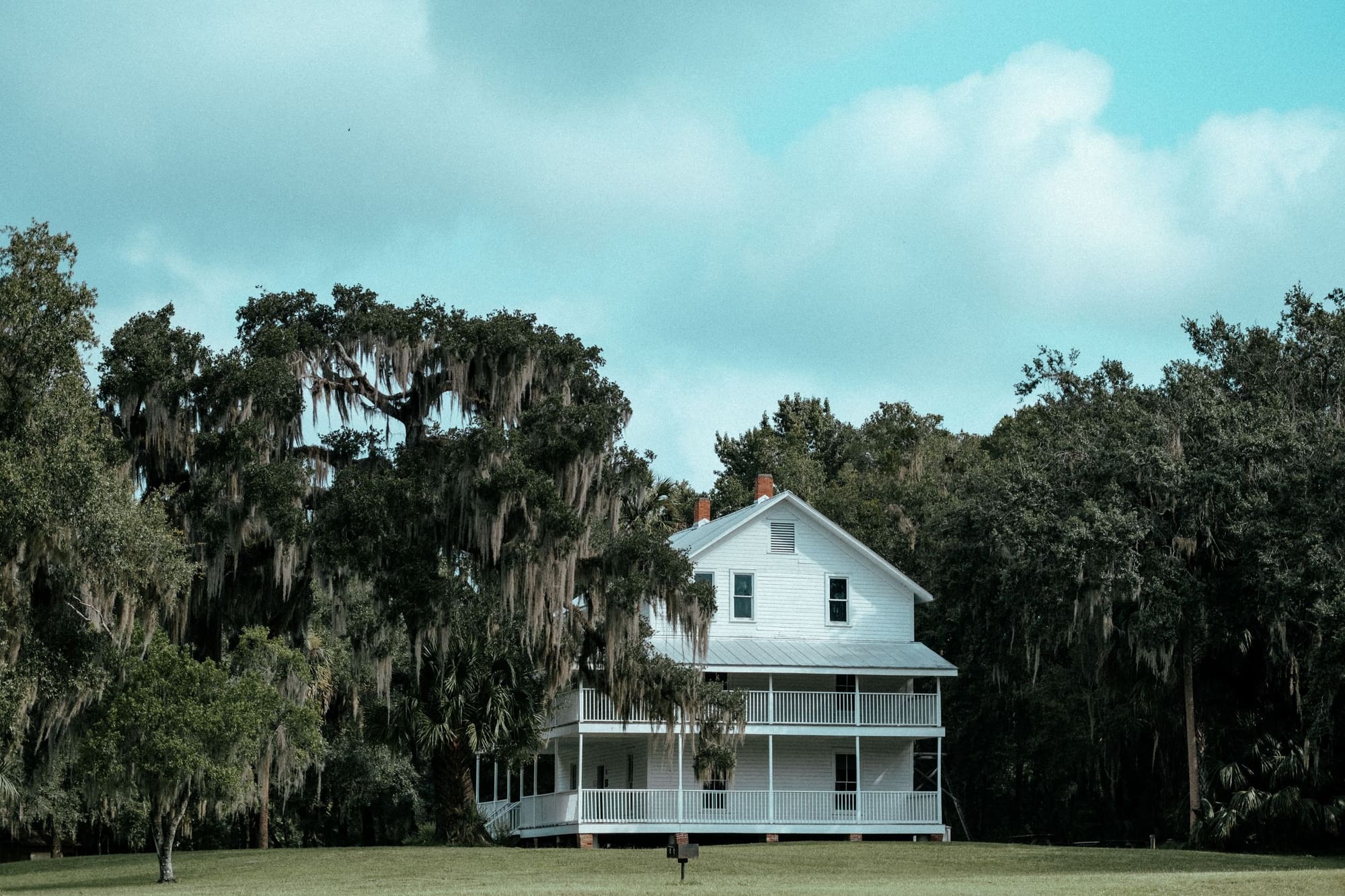 A white two-story farmhouse set back beneath tall live oaks draped in Spanish moss