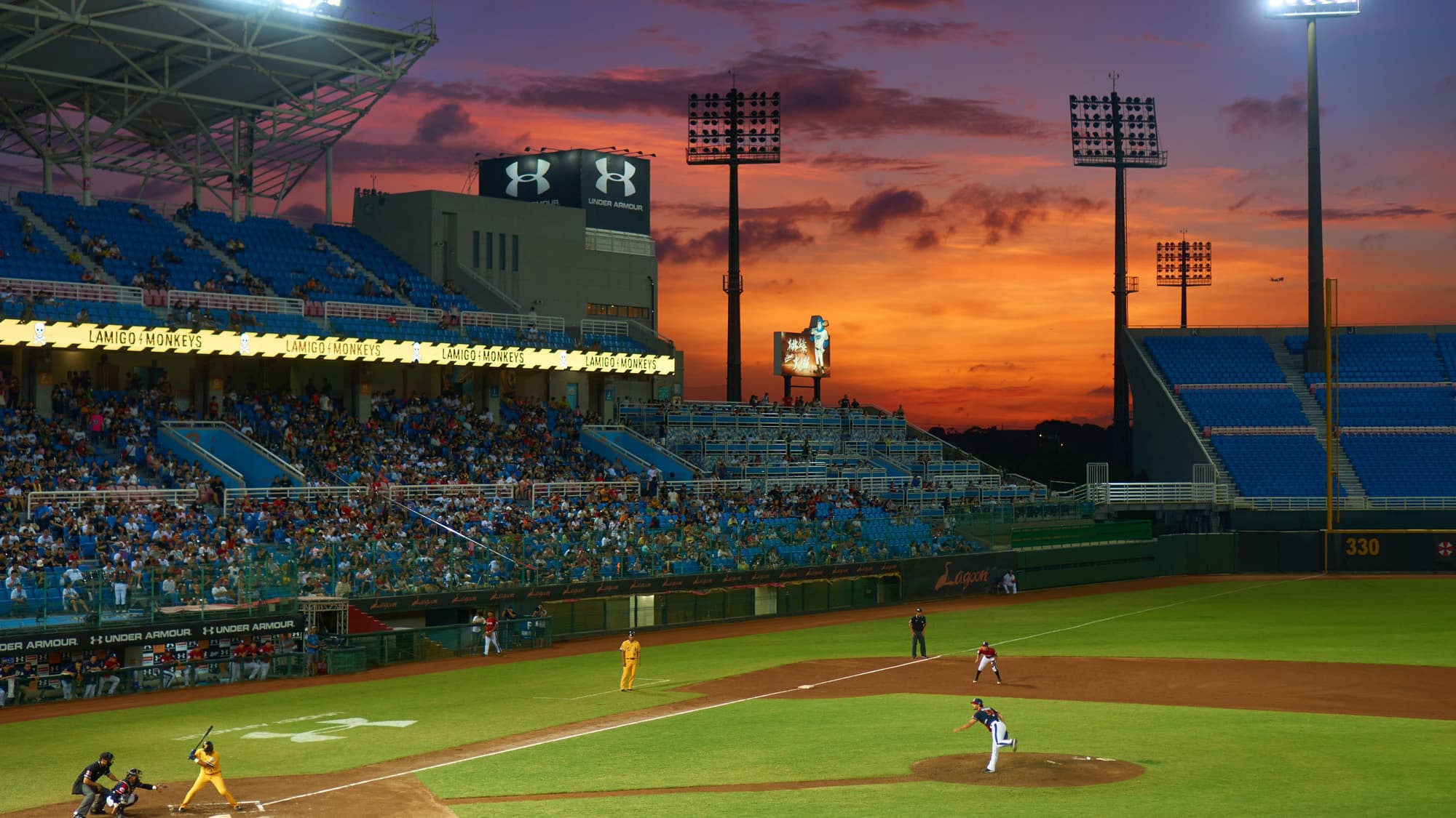 A baseball game under the stadium lights at dusk, with an orange and purple sunset over the stands